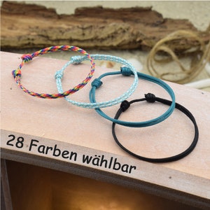 May include: Three colorful friendship bracelets made of woven string. The bracelets are in rainbow, light blue, and black colors. The text "28 Farben w&auml;hlbar" is visible on the wooden surface.