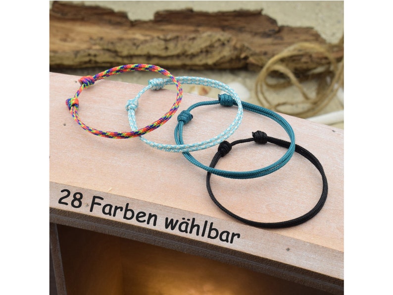 May include: Three colorful friendship bracelets made of woven string. The bracelets are in rainbow, light blue, and black colors. The text "28 Farben w&auml;hlbar" is visible on the wooden surface.