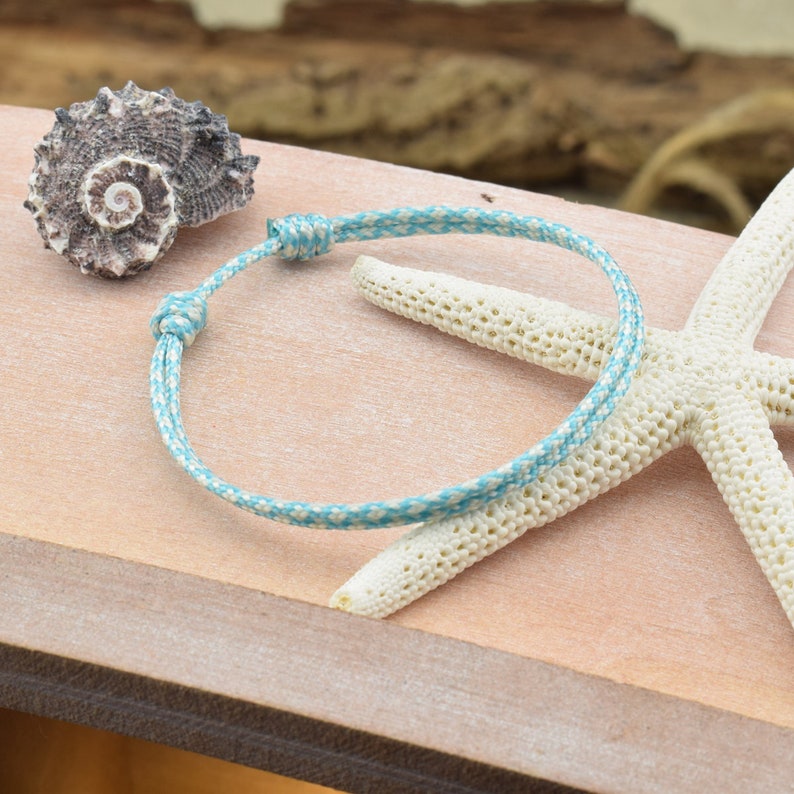 May include: A blue and white braided bracelet with a knot closure. The bracelet is photographed on a wooden surface with a seashell and a starfish.