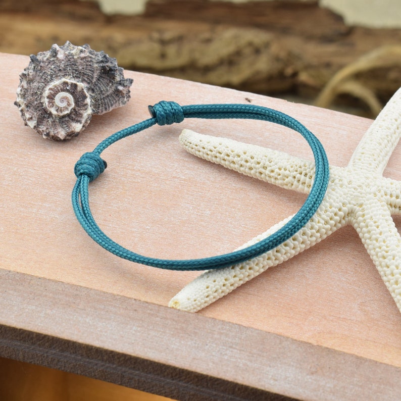 May include: A teal blue braided cord bracelet with a knotted closure. The bracelet is photographed on a wooden surface with a seashell and a starfish.