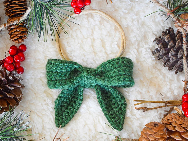 May include: A green crocheted bow with a wooden ring, surrounded by pine branches, pine cones, and red berries.
