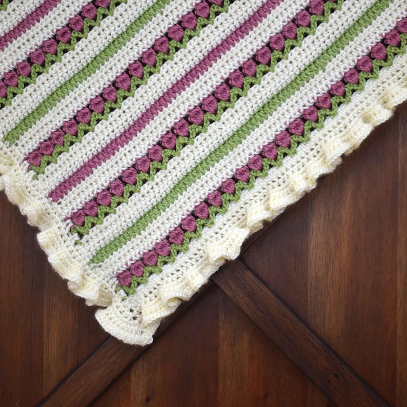 May include: A crocheted baby blanket with a white, green, and pink striped pattern. The blanket has a scalloped edge and is made with a delicate stitch.