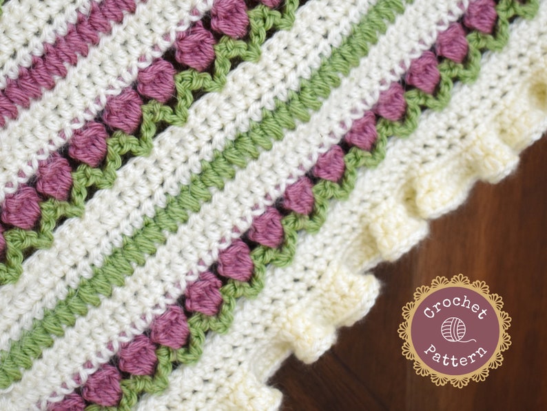 May include: Close-up of a crocheted blanket with a striped pattern in white, green, and pink. The blanket has a scalloped edge and features a repeating flower design.