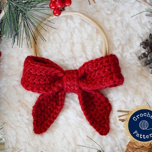 May include: A red crocheted bow tied around a beige headband. The bow is made with a chunky yarn and has a textured look. The headband is a simple elastic band.