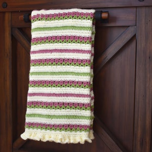 May include: A crocheted baby blanket with a striped pattern in shades of pink, green, and white. The blanket has a ruffled edge and is hanging on a wooden door.