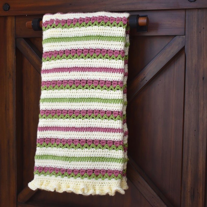 May include: A crocheted baby blanket with a striped pattern in shades of pink, green, and white. The blanket has a ruffled edge and is hanging on a wooden door.