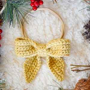 May include: A yellow crocheted bow tied around a brown elastic headband. The headband is surrounded by pine branches, pine cones, and red berries.