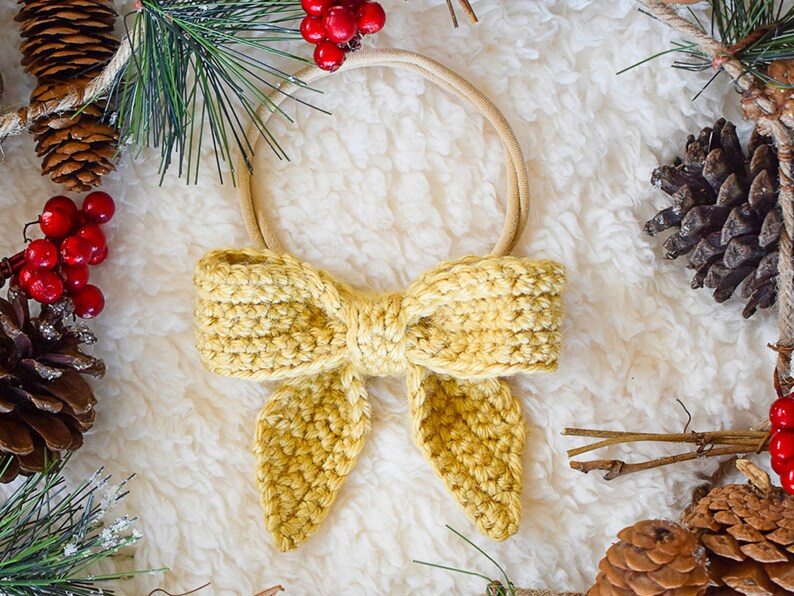 May include: A yellow crocheted bow tied around a brown elastic headband. The headband is surrounded by pine branches, pine cones, and red berries.