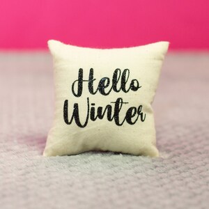 May include: A white throw pillow with the text "Hello Winter" in black script font.