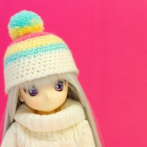 May include: A doll with long gray hair and purple eyes wearing a white sweater and a white crocheted hat with a rainbow stripe design and a pom-pom. The doll is holding a pink crocheted heart against a bright pink background.