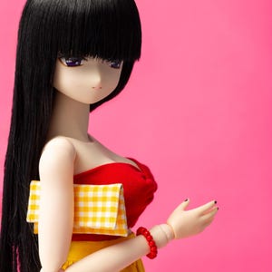 May include: A fashion doll with long black hair and purple eyes, wearing a red top and yellow skirt. The doll holds a yellow and white checkered clutch and a red beaded bracelet. The background is a solid pink.