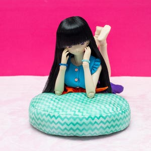 May include: A doll with long black hair wearing a blue and orange dress sits on a turquoise and white patterned cushion.