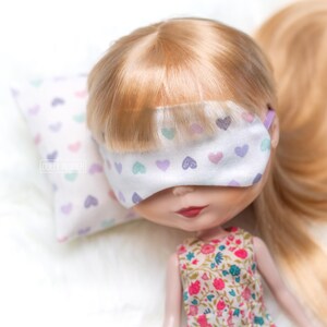 May include: A doll with blonde hair wearing a white sleep mask with a pink, purple and green heart pattern. The doll is lying on a white pillow with a pink, purple and green heart pattern.