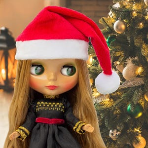 May include: A doll with long blonde hair and large green eyes wearing a red and white Santa hat and a black dress with gold trim. The doll is in front of a Christmas tree decorated with baubles.