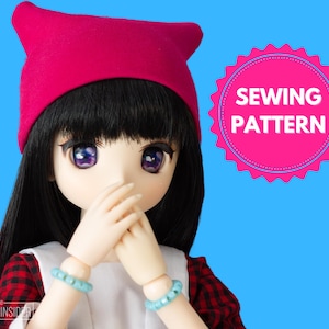 Puede incluir: Una muñeca con un sombrero rojo con orejas de gato, una camisa de cuadros rojos y negros y un delantal blanco. La muñeca está sosteniendo sus manos frente a su cara. La muñeca tiene el pelo largo y negro y ojos azules. La imagen tiene el texto "SEWING PATTERN" en un círculo rosa.
