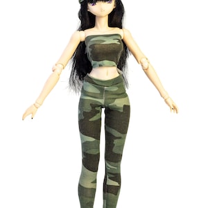 Puede incluir: Una muñeca con un atuendo de camuflaje verde, que incluye un top corto y leggings. La muñeca también lleva una boina verde y tiene el pelo largo y negro.