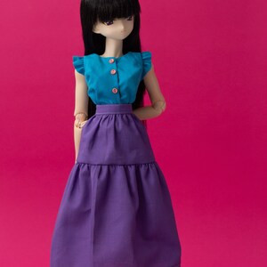 Puede incluir: Una muñeca con una blusa turquesa sin mangas con botones rosas y una falda de vuelo morada.