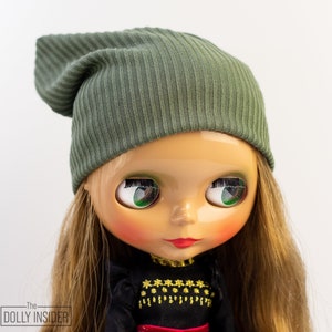 May include: A doll wearing a ribbed olive green beanie hat. The doll has long blonde hair, large green eyes, and red lipstick. The doll is wearing a black top with gold embellishments.