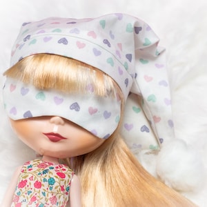 May include: A doll wearing a white sleep mask and cap, both patterned with pastel hearts. The doll has blonde hair and is wearing a floral print dress. The cap has a white pom-pom at the end.