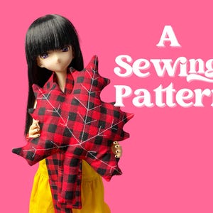 Puede incluir: Una muñeca sosteniendo una almohada y bufanda con forma de hoja a cuadros rojos y negros. El texto "A Sewing Pattern" se muestra en blanco, con un fondo rosa. La muñeca tiene cabello negro y viste un vestido amarillo.