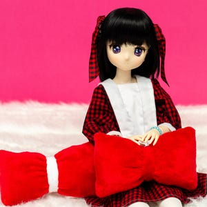 Peut inclure: Une poupée aux cheveux noirs et à la robe à carreaux rouges et noirs est assise sur une surface blanche et moelleuse. La poupée tient un coussin rouge en forme de nœud.