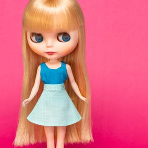 Könnte beinhalten: Puppe mit langem blondem Haar und blauen Augen, bekleidet mit einem blauen Oberteil und einem hellblauen Rock mit weißen Punkten. Der Hintergrund ist leuchtend rosa.