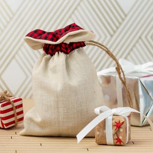 May include: A white fabric gift bag with a red and black plaid trim. The bag is tied with twine and is sitting on a wooden surface with three wrapped gifts. One gift is red and white striped, one is silver, and one is brown with white stars.