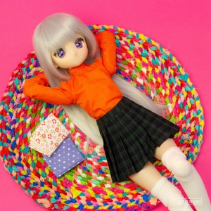 May include: A doll with silver hair and purple eyes wearing an orange shirt and a green plaid skirt, resting on a colorful braided rug. Two small books with floral and polka dot patterns are nearby. The background is a solid pink.