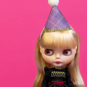 Puede incluir: Una muñeca con pelo largo rubio y ojos rosas que lleva un gorro de fiesta a cuadros morado con un pompón blanco y un ribete dorado con purpurina. La muñeca viste un vestido negro con bordados dorados y un cinturón rojo, sobre un fondo rosa brillante.