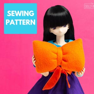 Peut inclure: Une poupée aux cheveux noirs et au haut bleu tient un grand nœud orange. Le nœud a un ruban rouge. La poupée porte une robe violette. Le texte "SEWING PATTERN" est dans un carré bleu en haut à gauche.