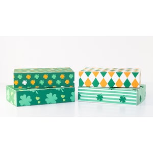 Puede incluir: Cuatro cajas rectangulares con diseños del Día de San Patricio. Dos cajas presentan tréboles verdes y blancos, mientras que las otras dos tienen patrones de diamantes y rayas en verde, blanco y naranja. Las cajas están apiladas en pares.