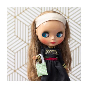 Könnte beinhalten: Eine Blythe-Puppe mit langem, hellbraunem Haar, blauen Augen und einem Stirnband. Die Puppe trägt ein schwarzes Kleid mit goldenen Verzierungen und einem roten Gürtel und hält eine kleine grüne Tragetasche mit weißen Punkten. Der Hintergrund zeigt ein gold-weißes geometrisches Muster.