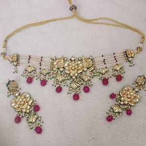 May include: A gold-toned necklace and matching earrings set. The necklace features a central floral design with white pearls, red beads, and green accents. The earrings mirror the necklace's design, with dangling red beads.