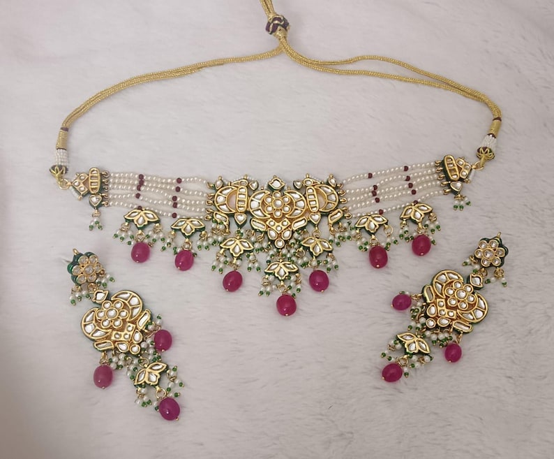 May include: A gold-toned necklace and matching earrings set. The necklace features a central floral design with white pearls, red beads, and green accents. The earrings mirror the necklace's design, with dangling red beads.