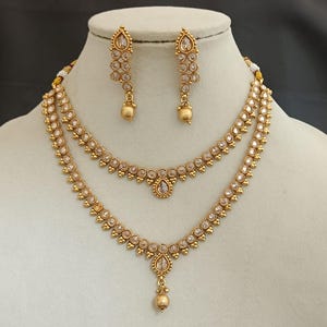 May include: A gold-toned jewelry set featuring a two-strand necklace and matching earrings. The necklace has a teardrop pendant and is adorned with clear, round gemstones. The earrings are a dangling style with similar gemstones and a small ball at the bottom.