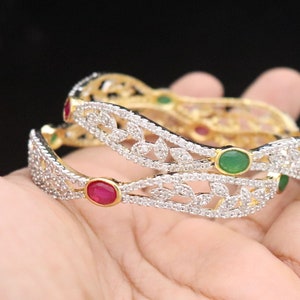 May include: Two gold bangles with a white diamond-like design and red and green gemstones.