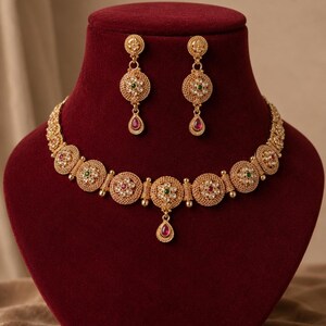 May include: A gold-toned necklace and earring set featuring circular filigree medallions. The necklace has a central teardrop pendant, while the earrings have matching drops. Each piece is accented with small white stones and central green and pink gems.