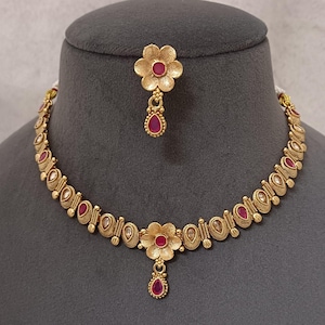May include: A gold-toned necklace and earring set. The necklace features a series of oval-shaped links with red and clear accents. The pendant and earrings have a flower design with a red center stone and a teardrop-shaped pendant.