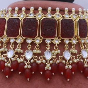 May include: A gold choker necklace with red and white beads. The necklace features a row of carved red stones with gold accents and a dangling row of red and white beads.