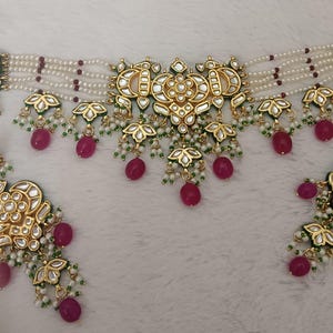 May include: A gold-toned necklace and earring set. The necklace features multiple strands of white beads with red accents, and gold pendants with green and white details. The earrings mirror the necklace design, with dangling red beads.