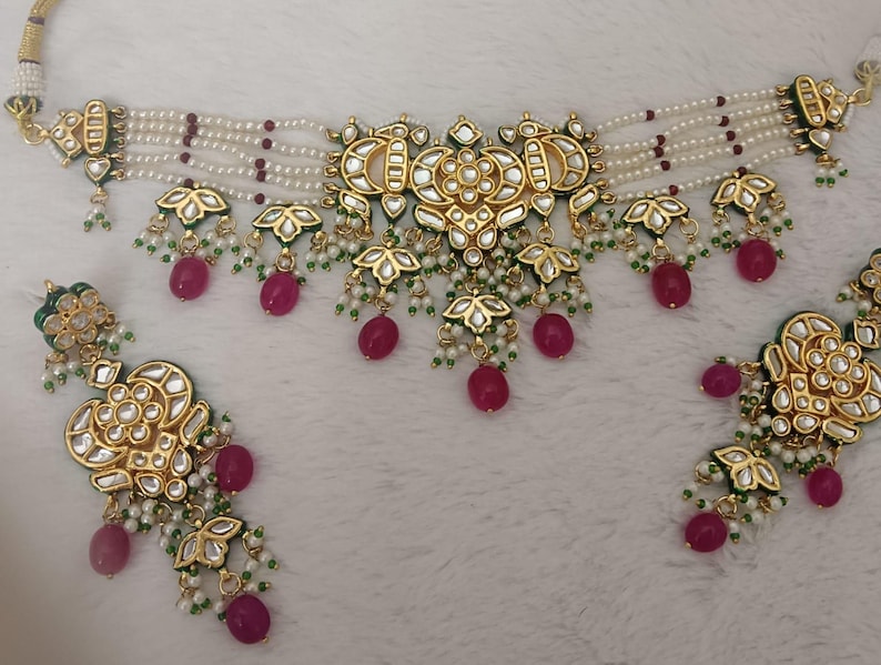 May include: A gold-toned necklace and earring set. The necklace features multiple strands of white beads with red accents, and gold pendants with green and white details. The earrings mirror the necklace design, with dangling red beads.