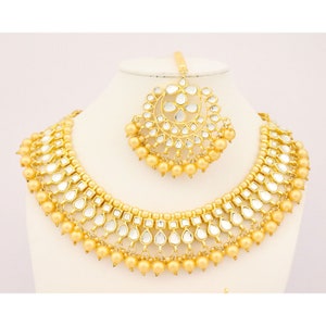 May include: A gold necklace and matching headpiece with a traditional Indian design. The necklace features multiple layers of gold beads and mirrored accents. The headpiece is a circular design with mirrored accents.