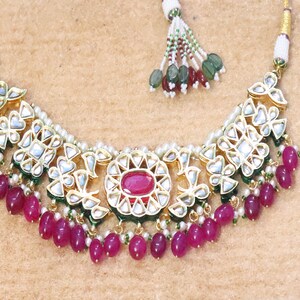 Pode incluir: Um colar de ouro com um grande pingente adornado com uma pedra preciosa vermelha cercada por pedras brancas menores. O colar é acentuado com contas rosa e verdes. Brincos combinando com um design semelhante também estão incluídos.