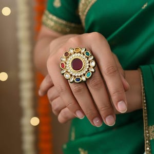 May include: A gold-toned statement ring featuring a large, round, deep red gemstone at its center, surrounded by smaller, colorful gemstones in red, white, yellow, blue, and green. The ring has an ornate, floral-like design, and is set against a green background.