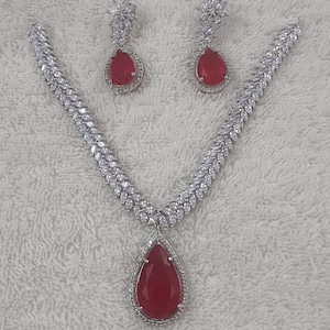 Op de afbeelding: Een zilveren ketting en oorbellen set met een traanvormige rode stenen hanger en bijpassende oorbellen. De ketting en oorbellen zijn versierd met heldere zirkonia stenen.