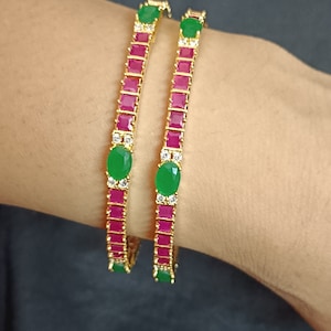 Peut inclure: Deux bracelets en or avec des pierres roses et vertes. Les bracelets sont décorés d'une rangée de pierres roses carrées et d'une pierre verte ovale plus grande à chaque extrémité.
