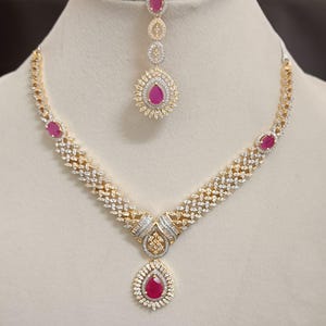 Op de afbeelding: Een goudkleurige ketting en hoofdband set. De ketting heeft een V-vormig ontwerp met heldere en rode edelstenen. De hoofdband heeft een bijpassende druppelvormige rode edelsteen hanger. De sieraden worden op een wit oppervlak getoond.