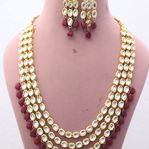 Puede incluir: Un collar de oro con cuatro hileras de piedras ovaladas y cuentas rojas. El collar se combina con pendientes a juego con cuentas rojas.