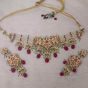 May include: An ornate necklace and earring set. The necklace features a central pendant with red, green, and white floral designs, accented with dangling red beads. The earrings mirror the necklace design. The set is strung on a gold-colored cord with white beads.