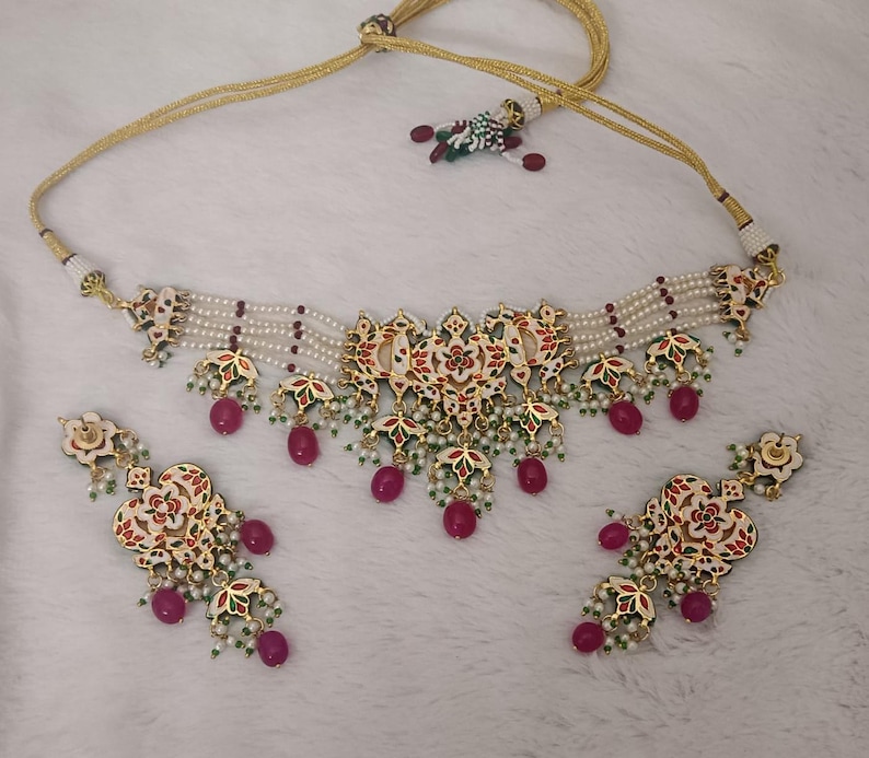 May include: An ornate necklace and earring set. The necklace features a central pendant with red, green, and white floral designs, accented with dangling red beads. The earrings mirror the necklace design. The set is strung on a gold-colored cord with white beads.